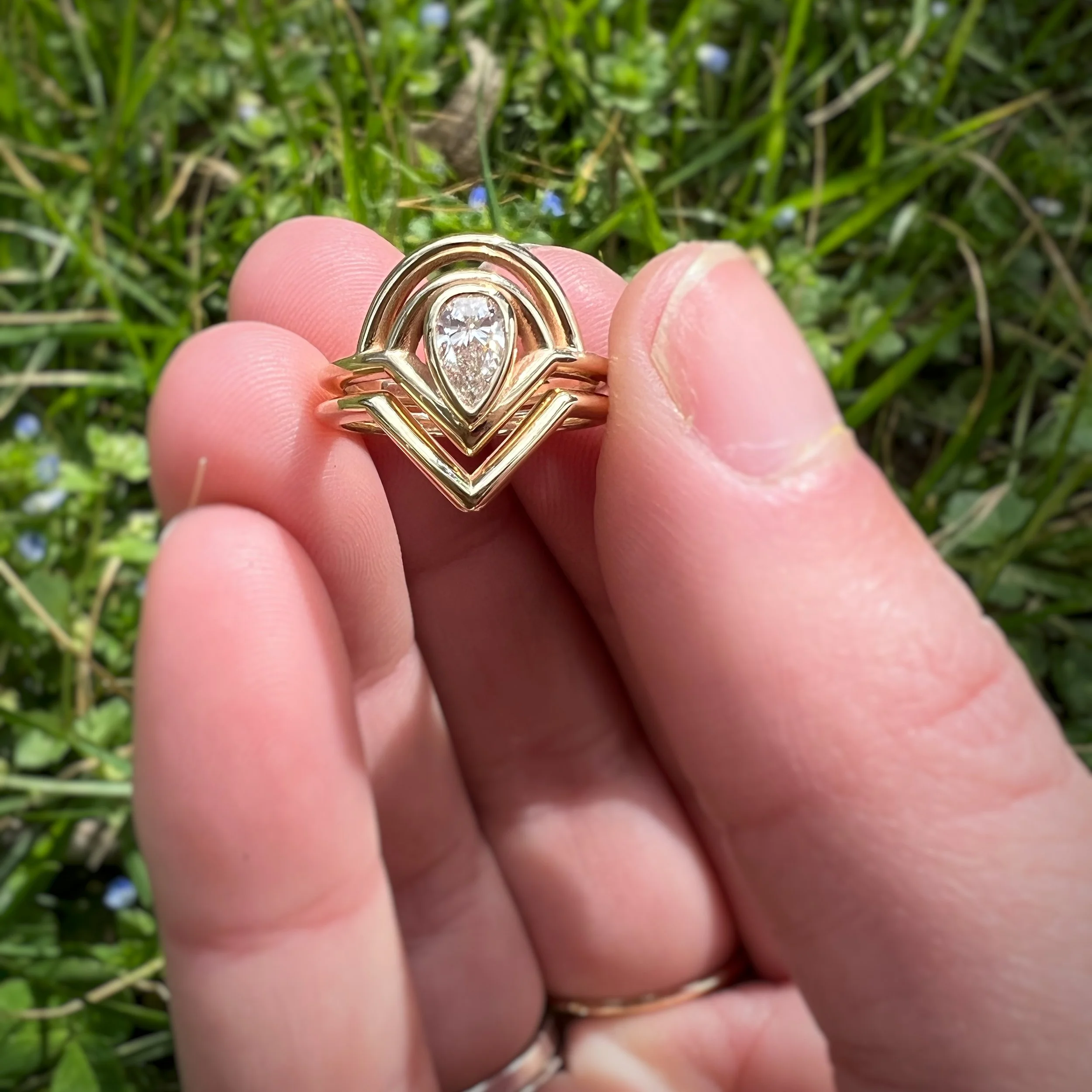 Close-up of a gold ring with an intricate design and a teardrop-shaped diamond in the center, held between fingers over green grass.