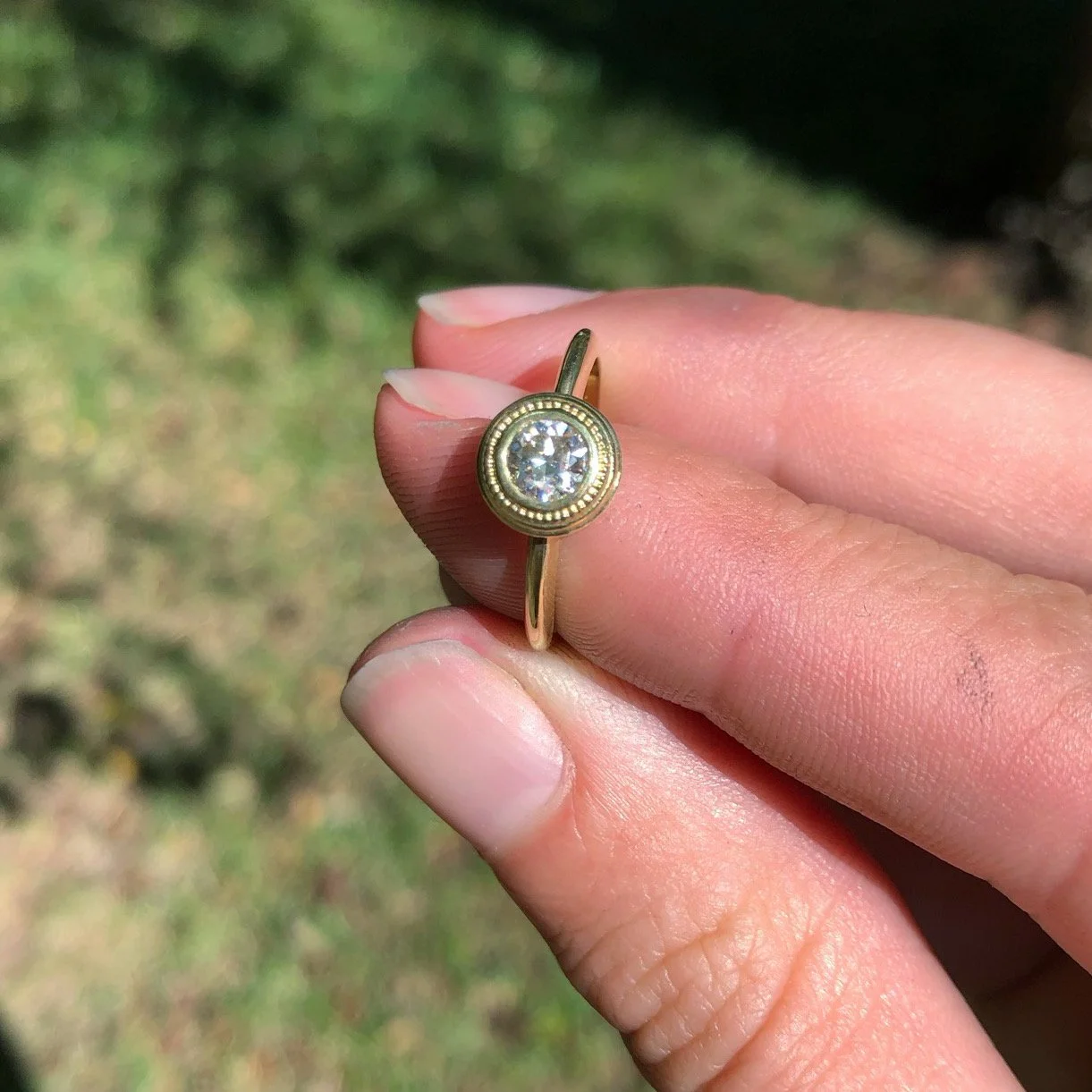 A gold ring with a large clear gemstone, held between the thumb and index finger of a person's hand, with a blurred green outdoor background.