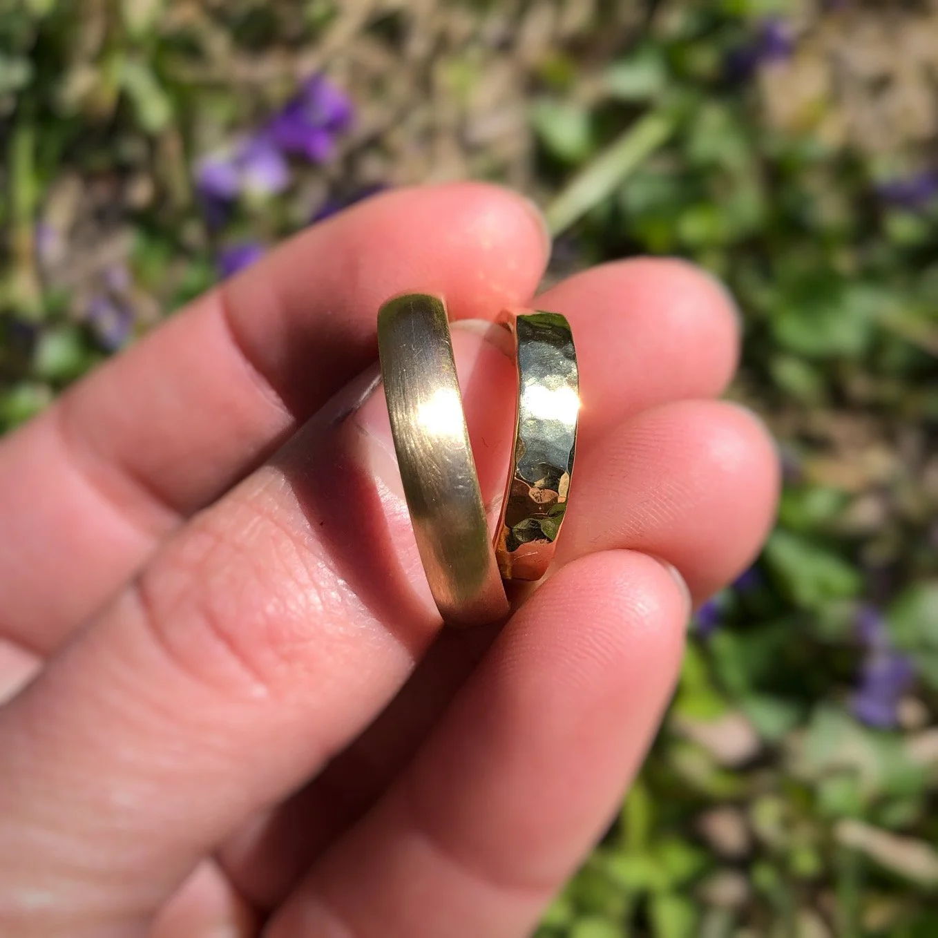 Two gold wedding rings held between fingers against blurred floral background.