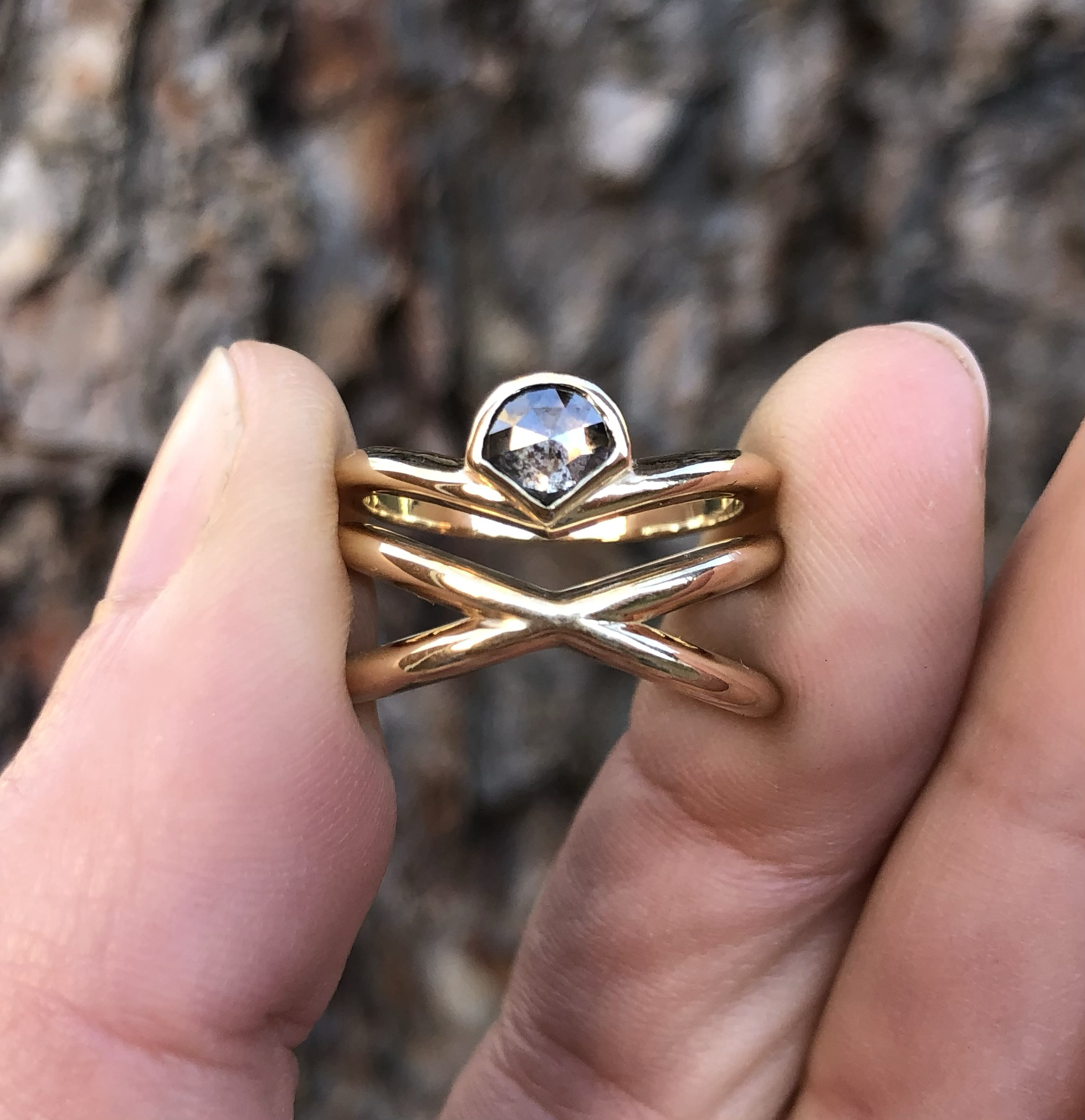 A gold ring with a diamond-shaped black gemstone, held between fingers against a blurred bark background.