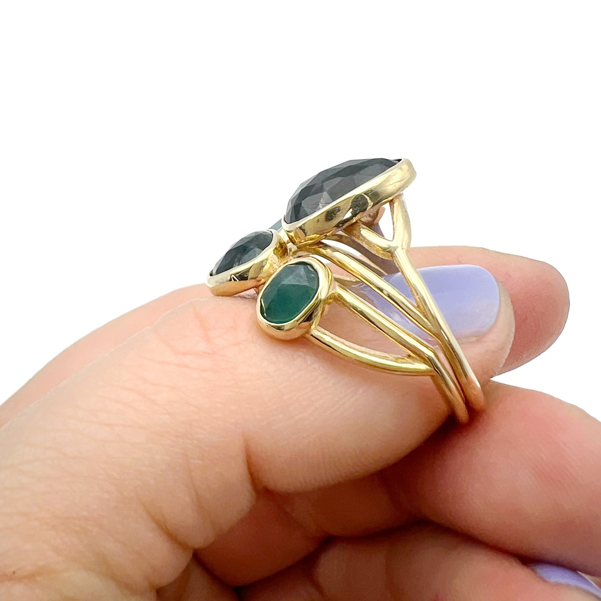 Gold ring with multiple dark-colored and green gemstones, held between fingers with light purple painted nails.