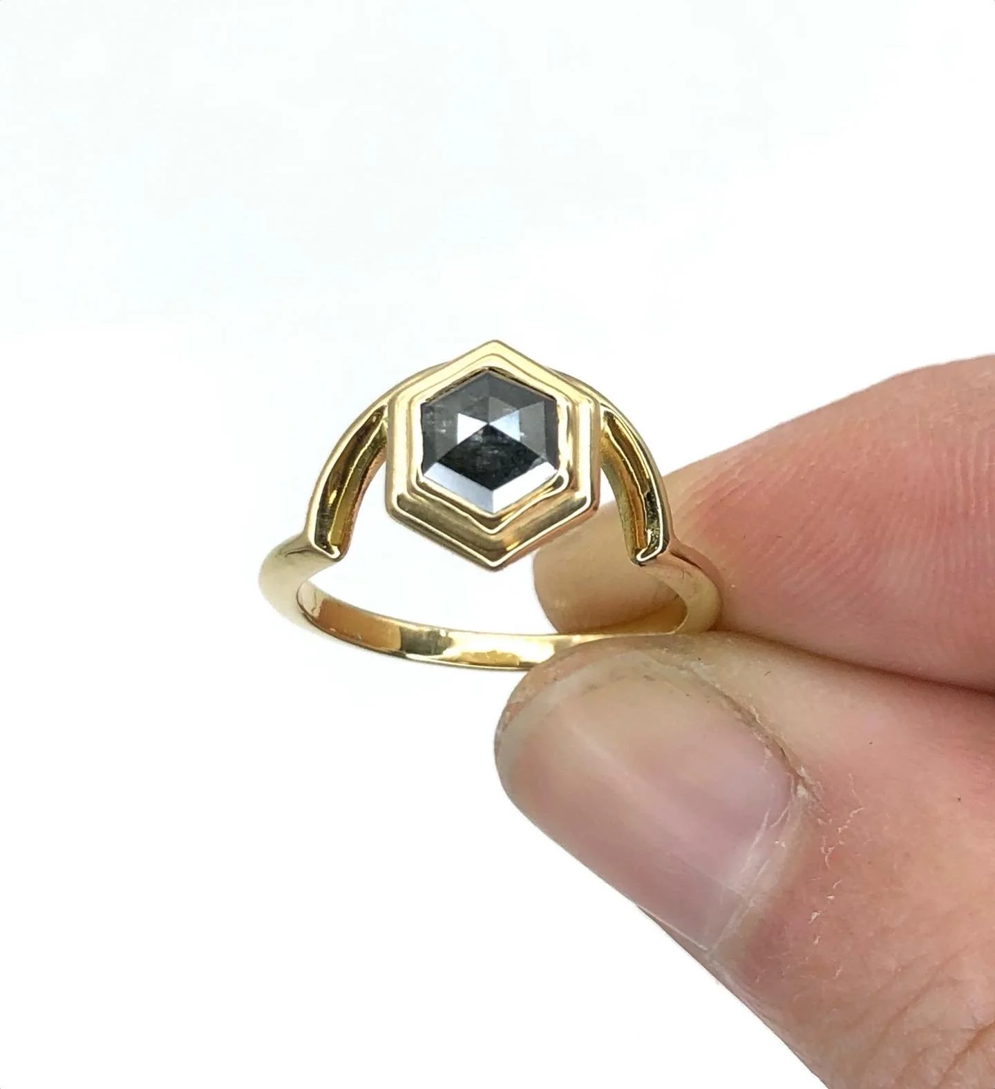 A gold ring with a hexagon-shaped black gemstone in the center, held between two fingers.