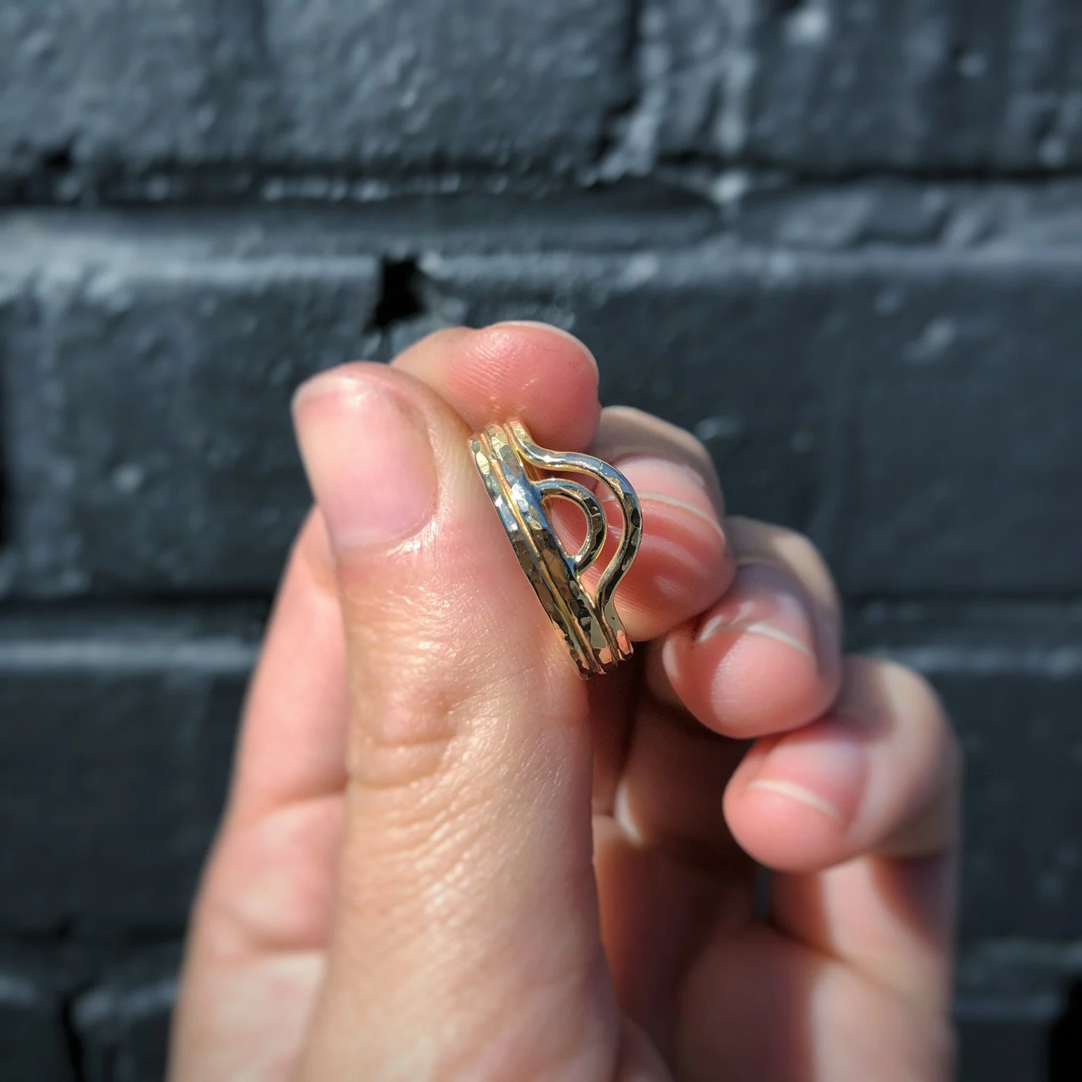 Close-up of a hand holding a gold ear cuff earring with a wavy design, with a dark textured wall in the background.
