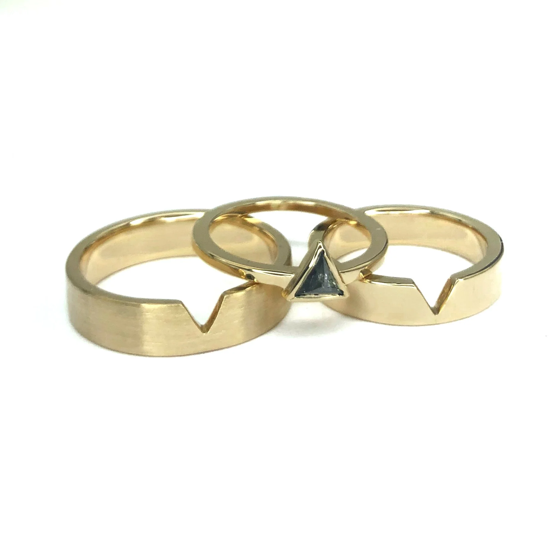 Three gold rings with geometric designs, two with cut-out shapes and one with a black triangular gemstone.