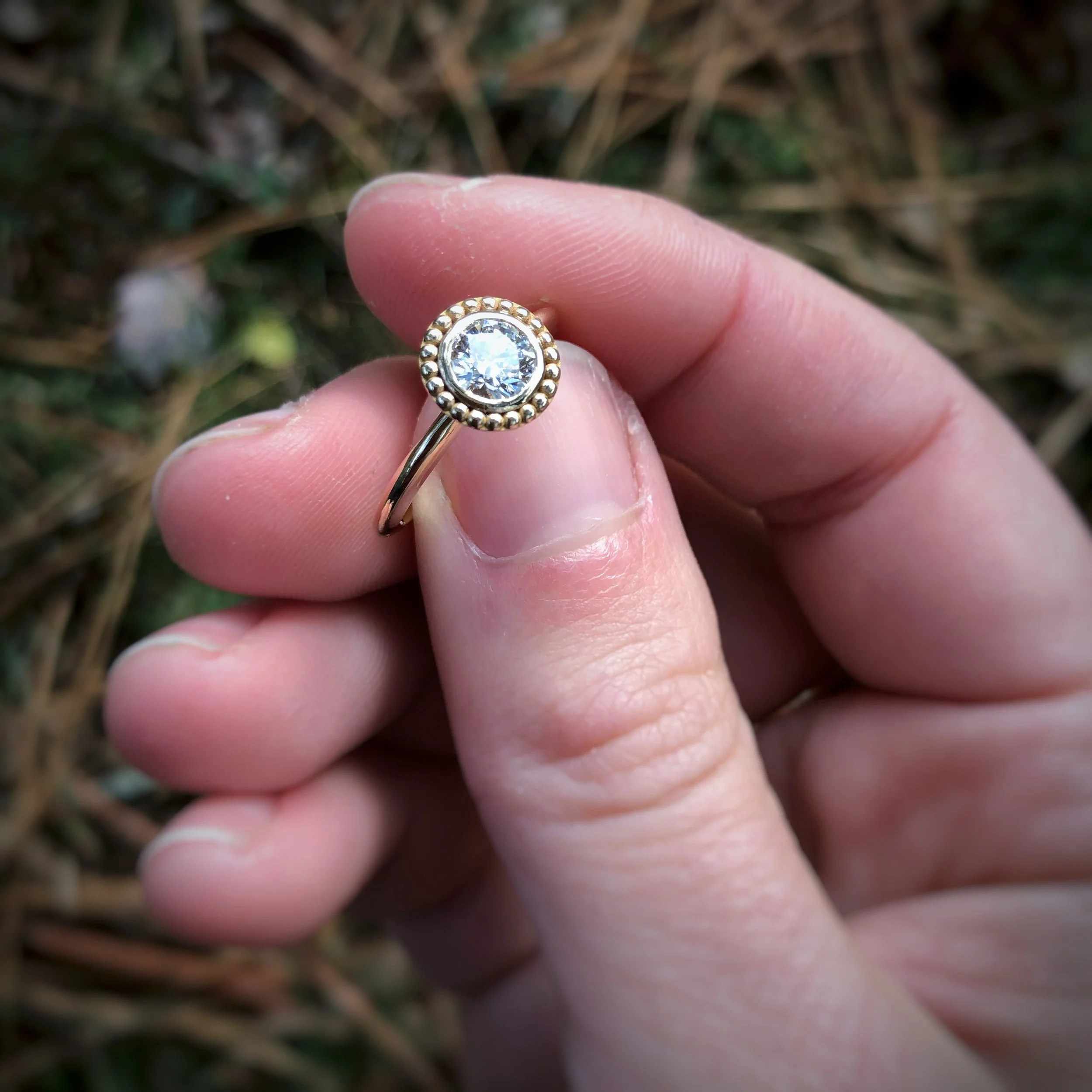 Close-up of a hand holding a gold ring with a round clear gemstone, surrounded by small beads, against a background of grass and dried leaves.