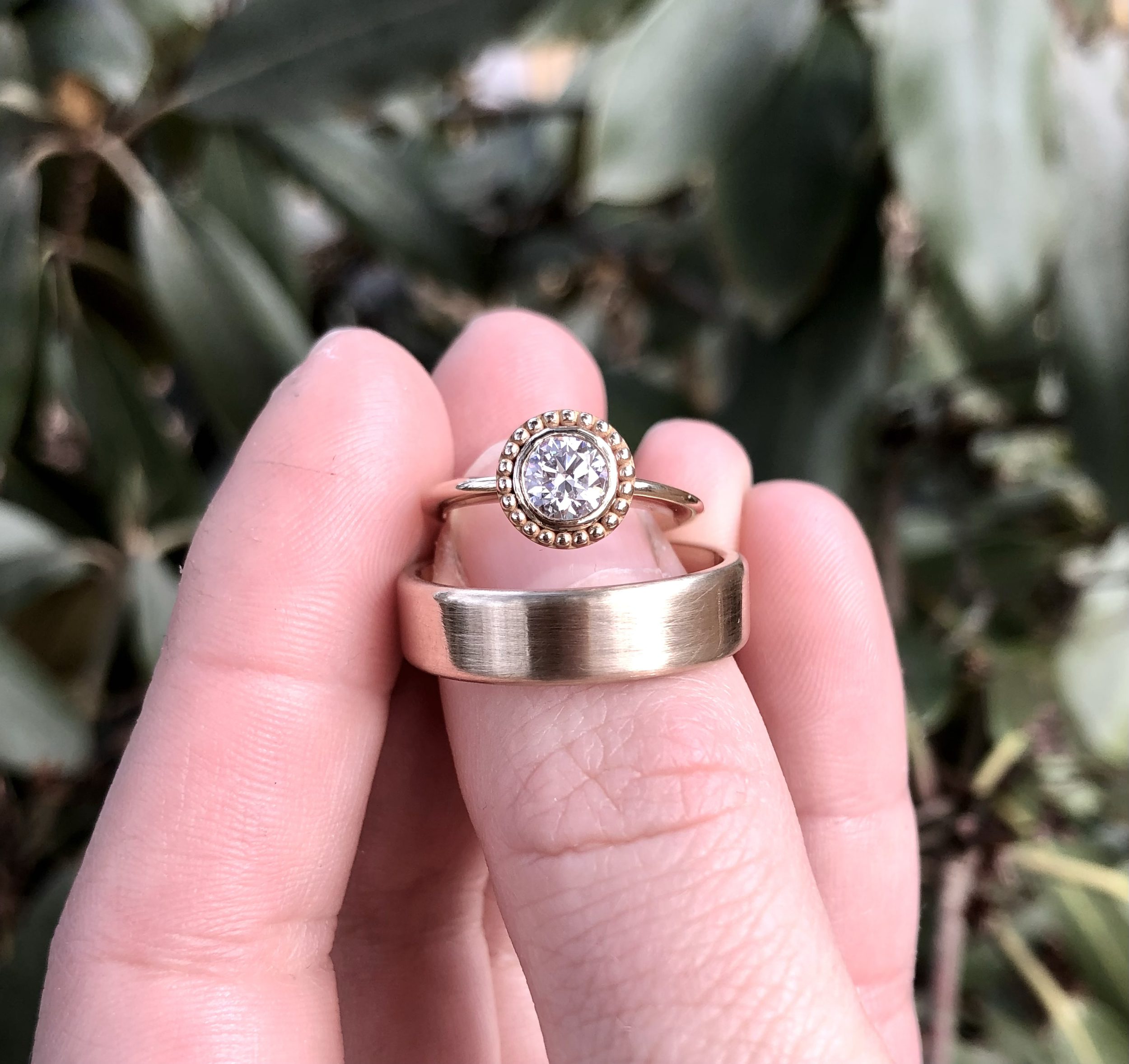 Two rings held between a person's fingers in front of a background of green leaves. One ring is a gold band with a brushed finish, the other a gold ring with a round diamond surrounded by smaller beaded details.