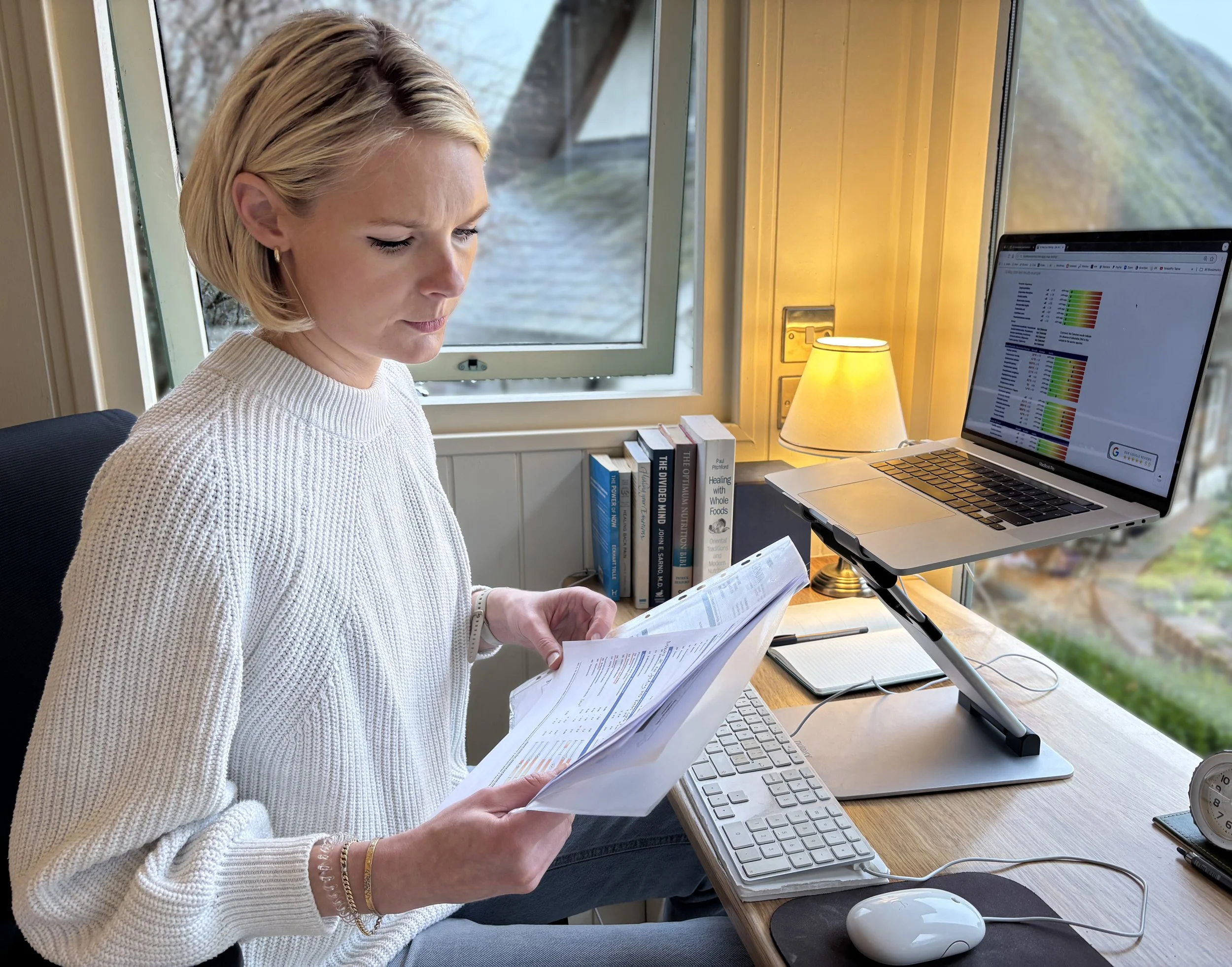 Georgie Spavins - nutritional therapist sitting at a desk, reading lab test reports, with a computer, books, and a lamp in a room with a window outside.