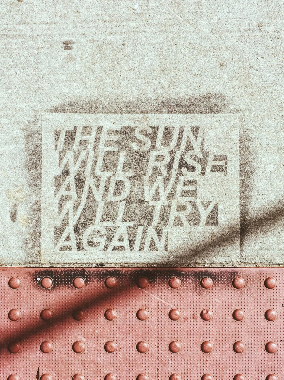 A concrete sidewalk with a stamped message that reads 'When the sun rises and we walk away, I say again.' The sidewalk has a section of textured red tactile paving with raised bumps for safety.