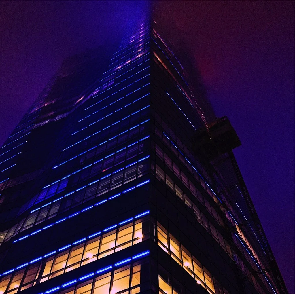 Nighttime photo of a tall modern skyscraper with glass windows outlined by blue and purple lights, with some illuminated indoor lights visible through the windows.