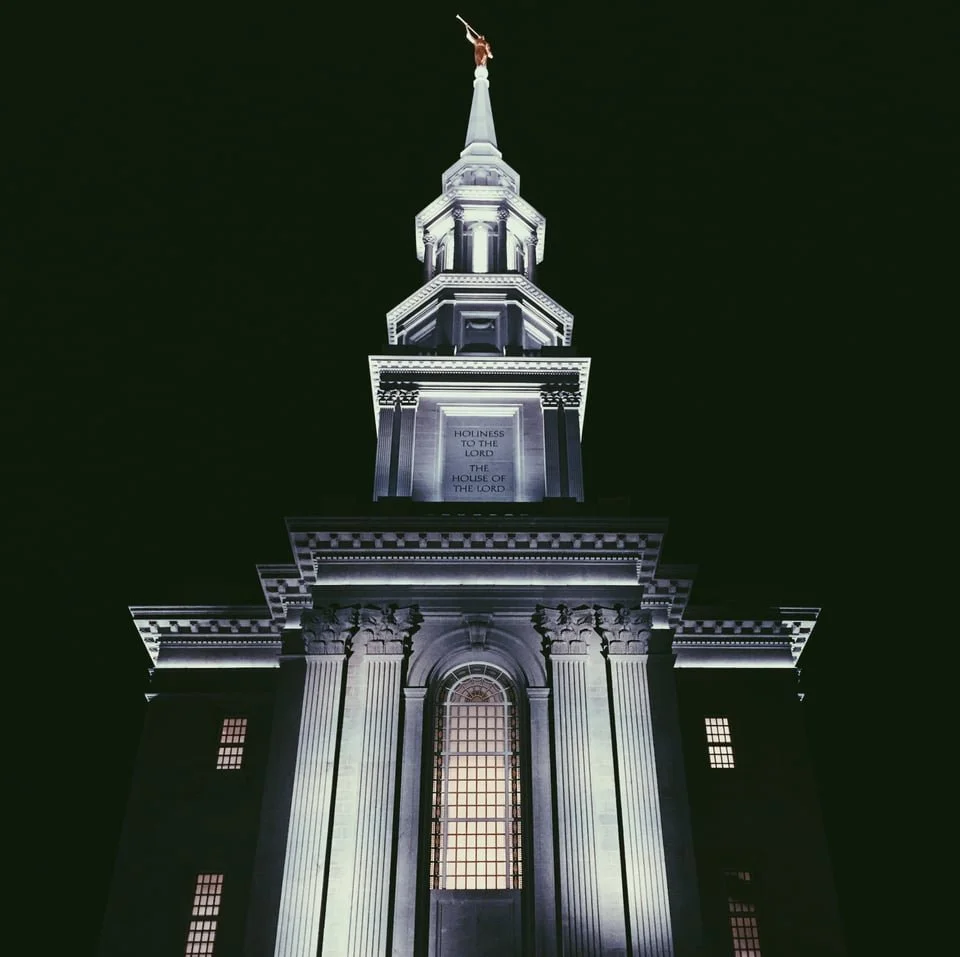 Night view of a tall, illuminated church steeple with a golden weather vane on top, featuring classical architectural details like columns and decorative moldings.