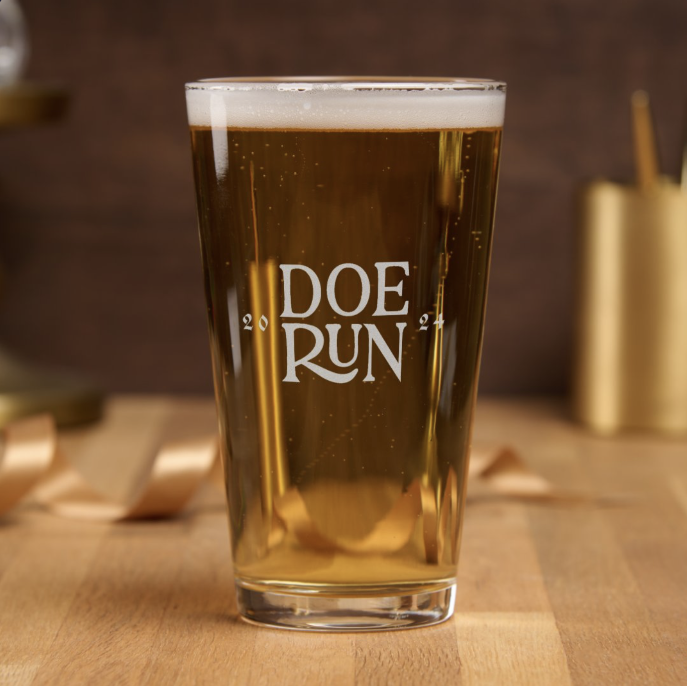 A pint glass filled with beer, with 'DOE RUN' logo printed on it, placed on a wooden surface with blurred background.