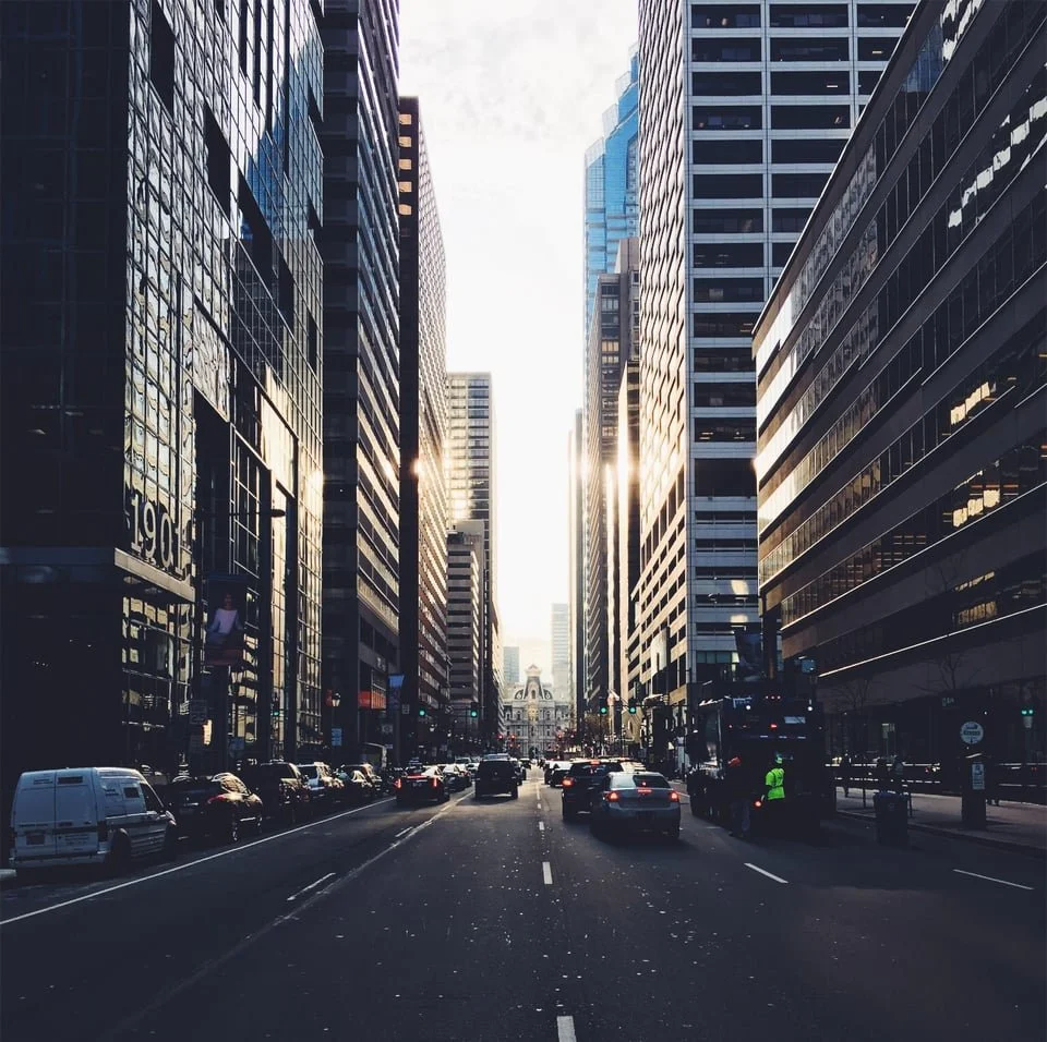 A busy city street lined with tall glass skyscrapers on both sides, with cars and a construction worker on the road, sunlight shining through the buildings.