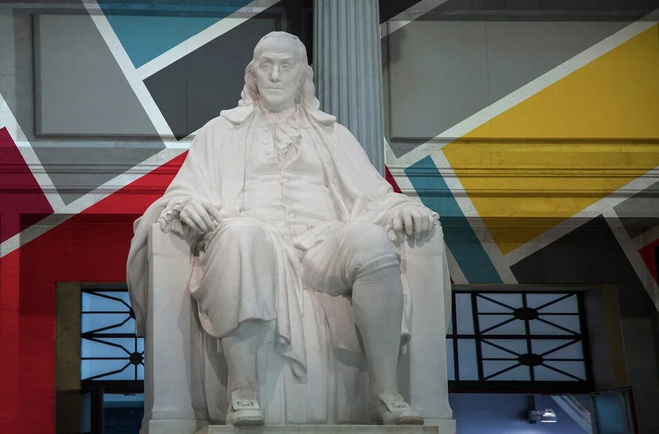 White marble statue of Benjamin Franklin sitting in a chair with colorful modern geometric wall art behind it.