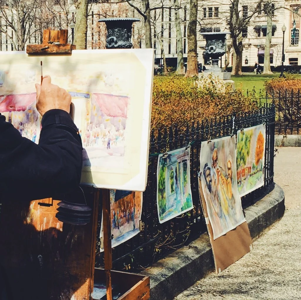 An artist painting watercolor scenes outdoors near a park with trees and ornate lampposts, with paintings of nature and people displayed on a black iron fence.