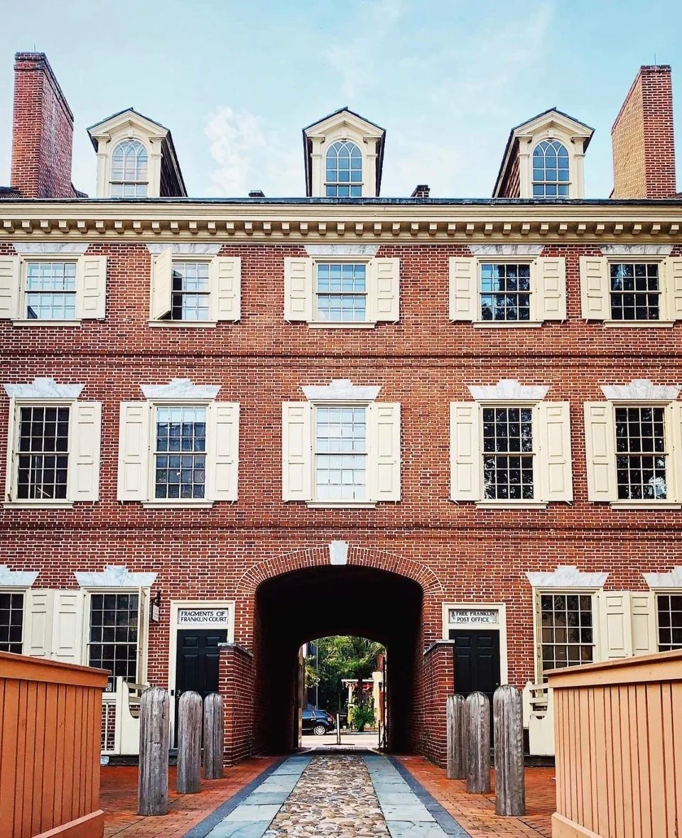 Red brick building with white window shutters and dormer windows on the roof, archway entrance with signs indicating fragments of Franklin Court and street directions, walkway with cobblestone path and wooden posts on either side.