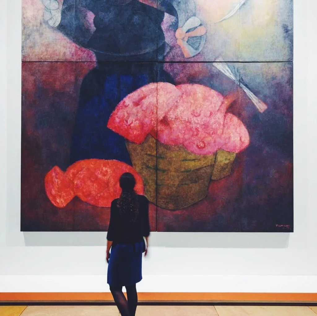 A woman stands in front of a large abstract painting of a person eating a cupcake with pink frosting, holding a spoon, in an art gallery.