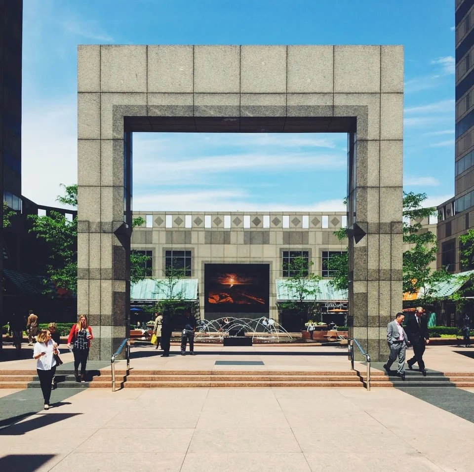 A city plaza with a large stone archway, a fountain, and people walking around, with office buildings and trees in the background.