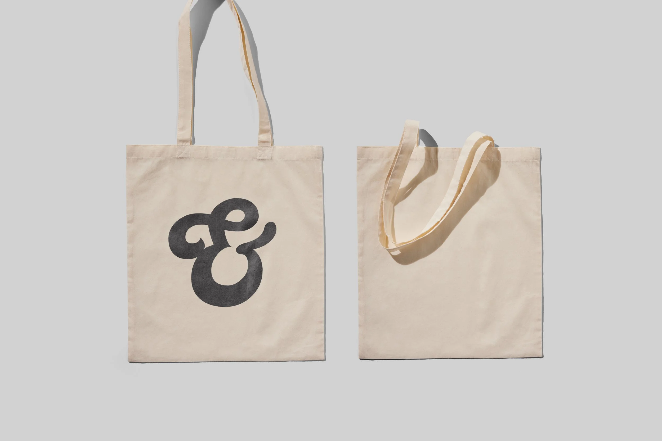 Two cream-colored tote bags on a light gray background, one with a black abstract design and the other plain.