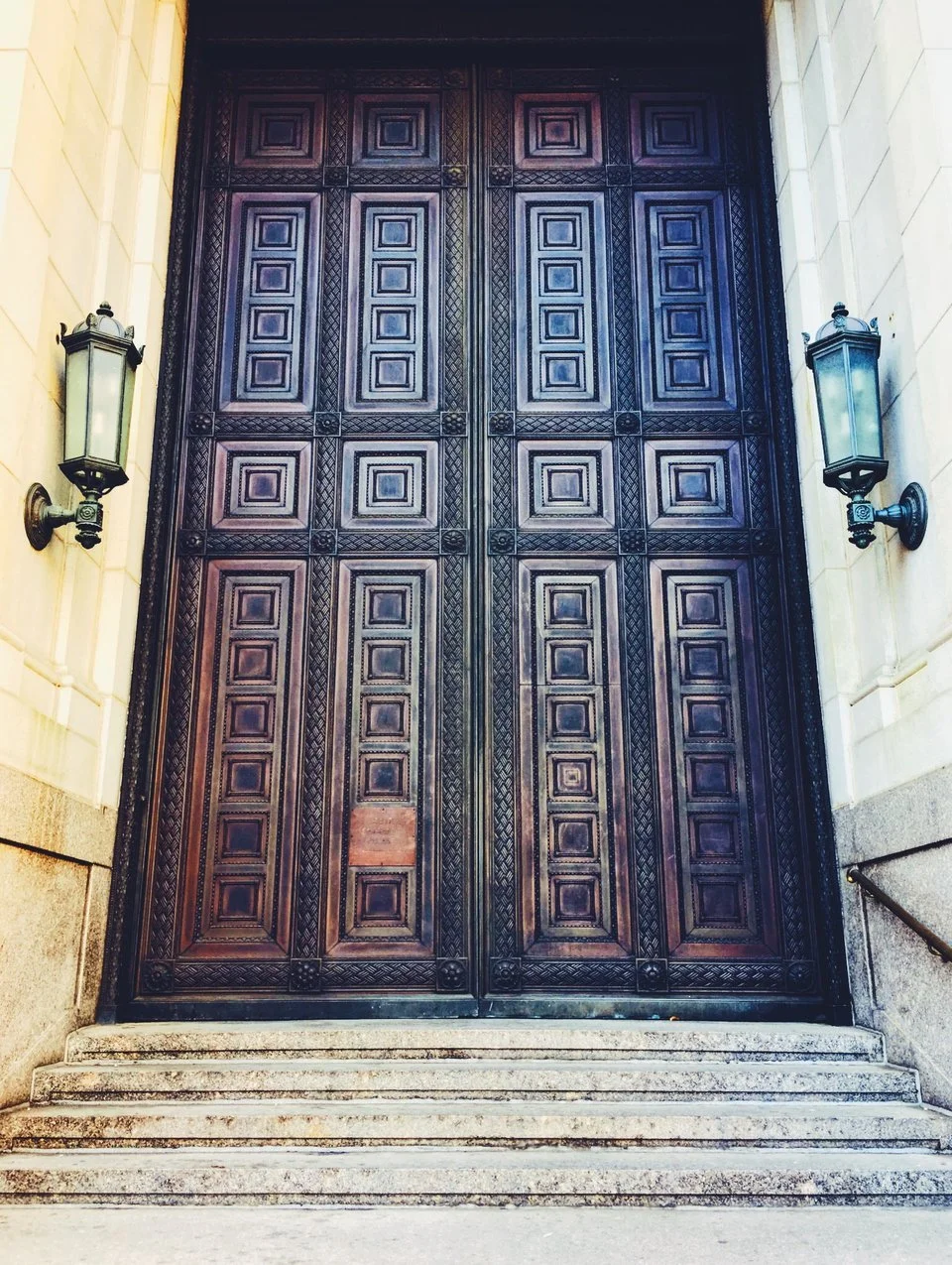 Large ornate wooden double door with geometric patterns, flanked by two wall-mounted lanterns, set at the top of a stone staircase.