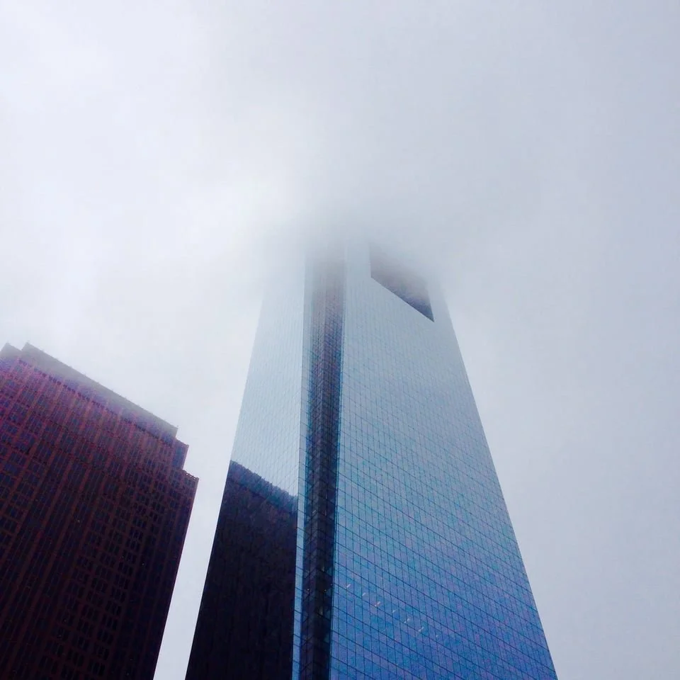A tall glass building partially covered by fog in a city skyline.