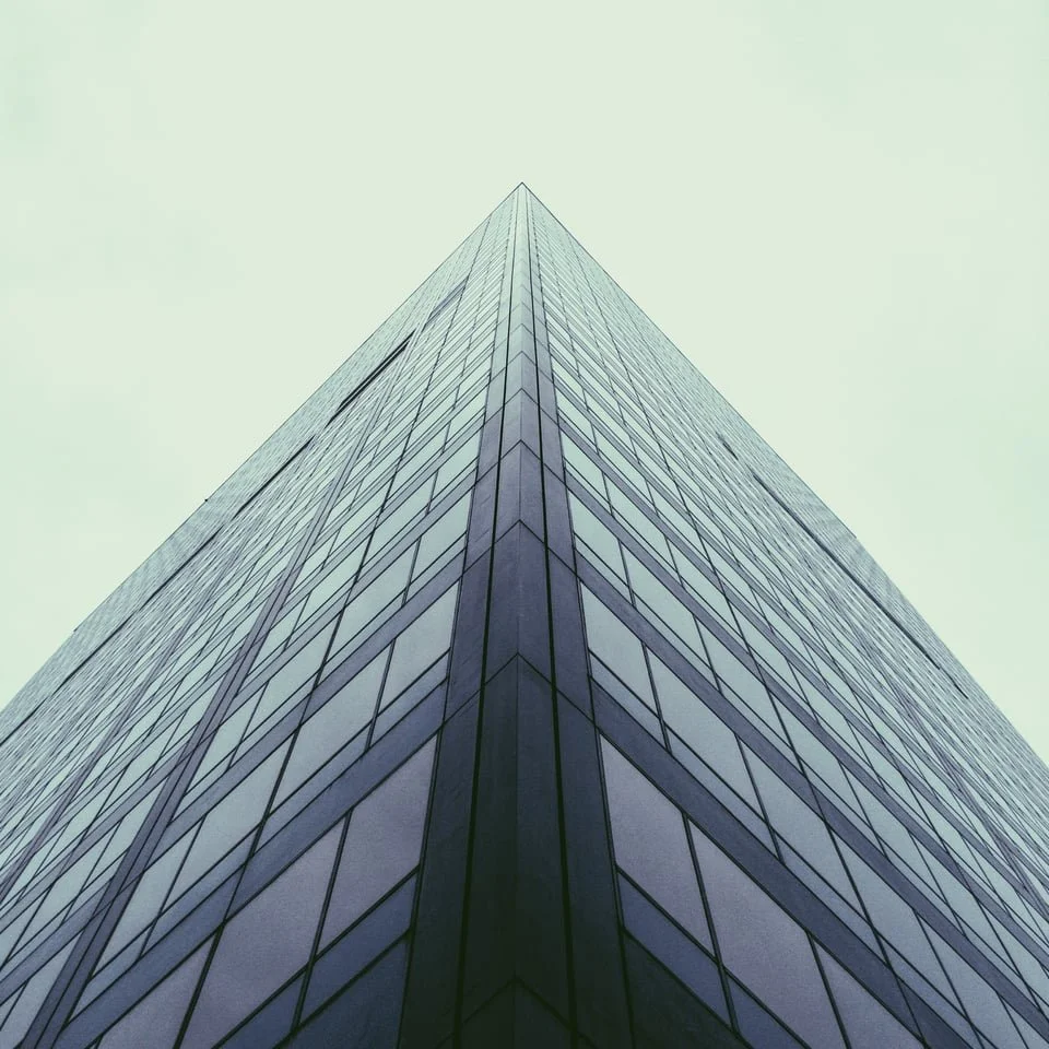 A tall glass skyscraper viewed from the ground looking up, with reflective windows and a cloudy, pale sky in the background.