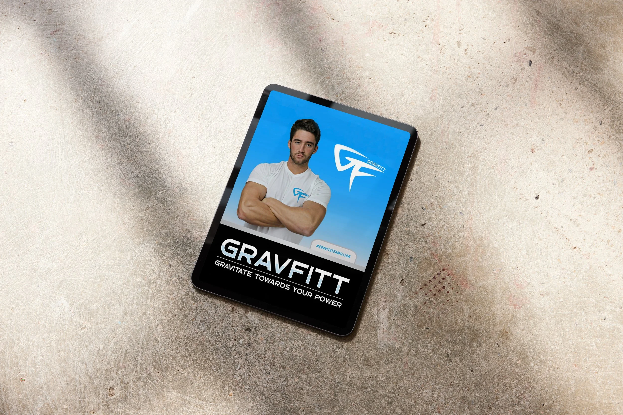 Smartphone on a textured surface displaying a fitness app with a photo of a muscular man crossing his arms, wearing a white T-shirt, with a blue background and the logo 'Gravitt' along with the caption 'Gravitate towards your power'.