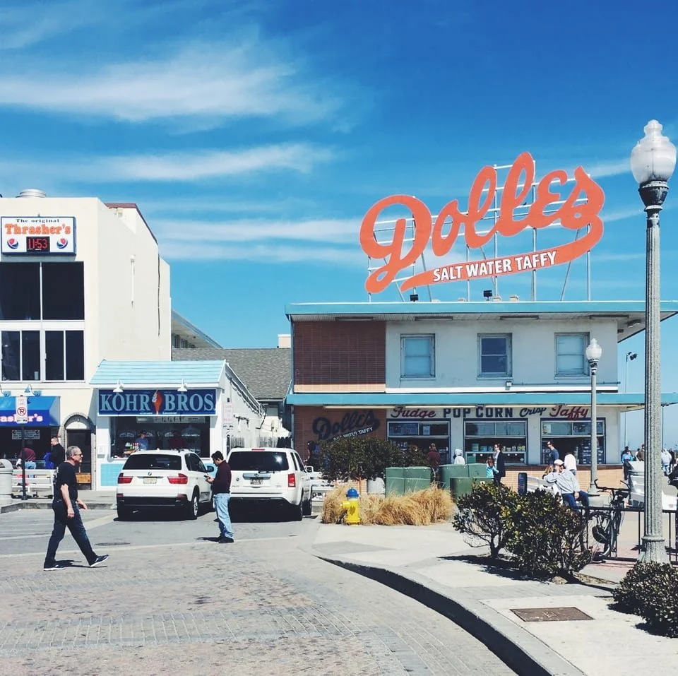 People walking on a street next to a building with a large sign that reads "Dolly's Salt Water Taffy," with a smaller sign for "Thrasher's" and a shop called "Kohler Brothers." Cars are parked in front of the shops, and there are street lamps and bus