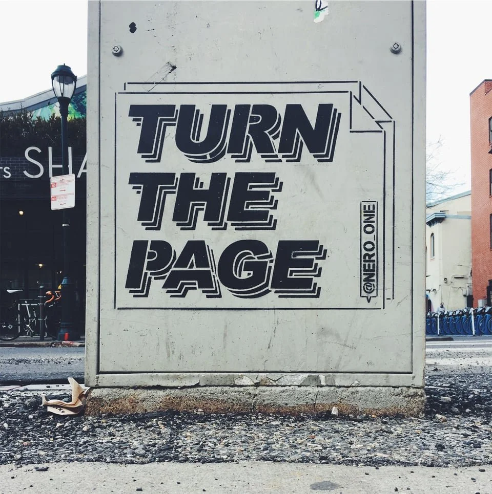Street art mural with the words "TURN THE PAGE" painted in bold black font on a gray utility box.