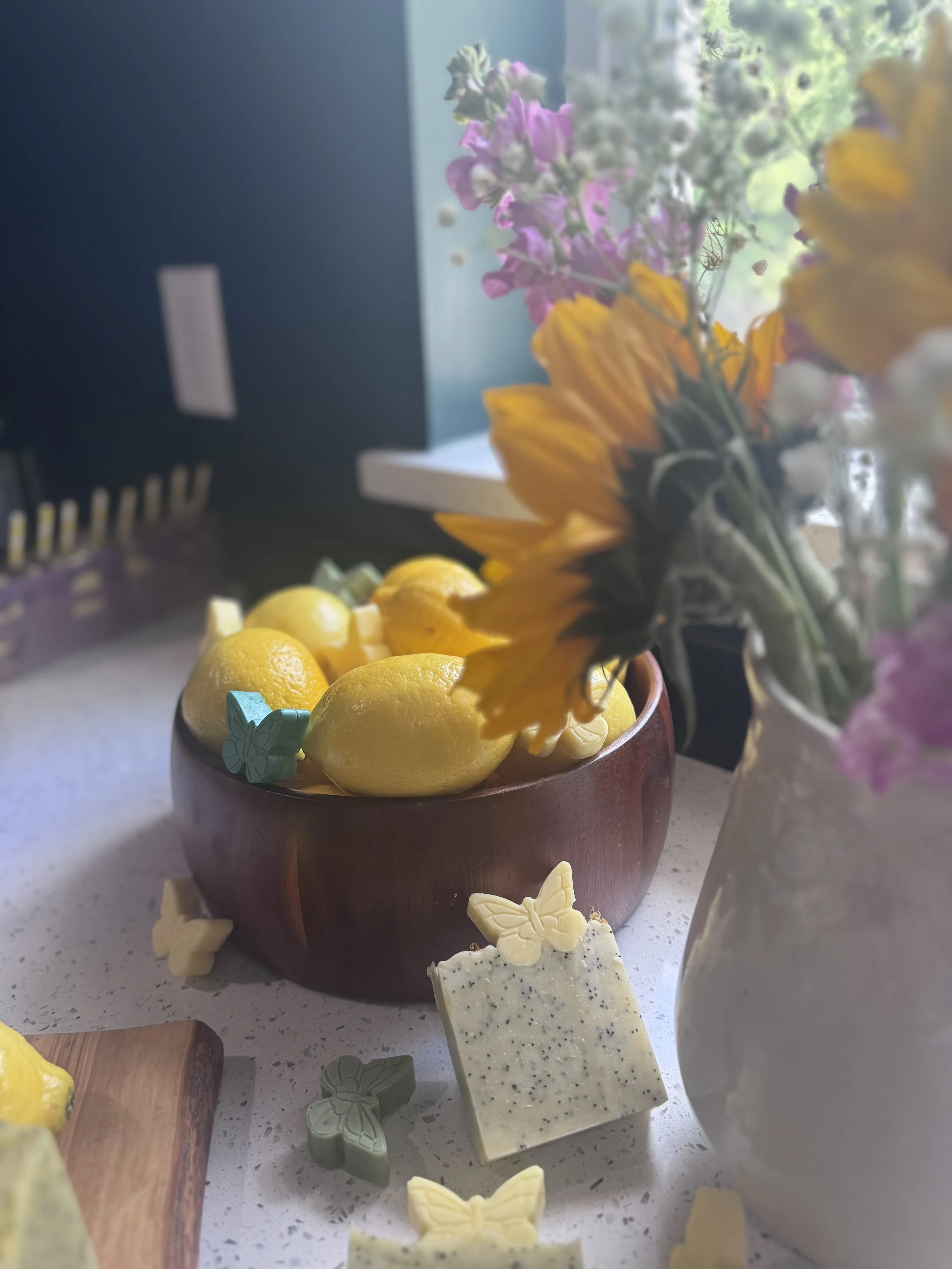 A kitchen countertop with a bowl of lemons, some lemon-themed soap, and a vase of colorful flowers, with a window in the background letting in natural light.