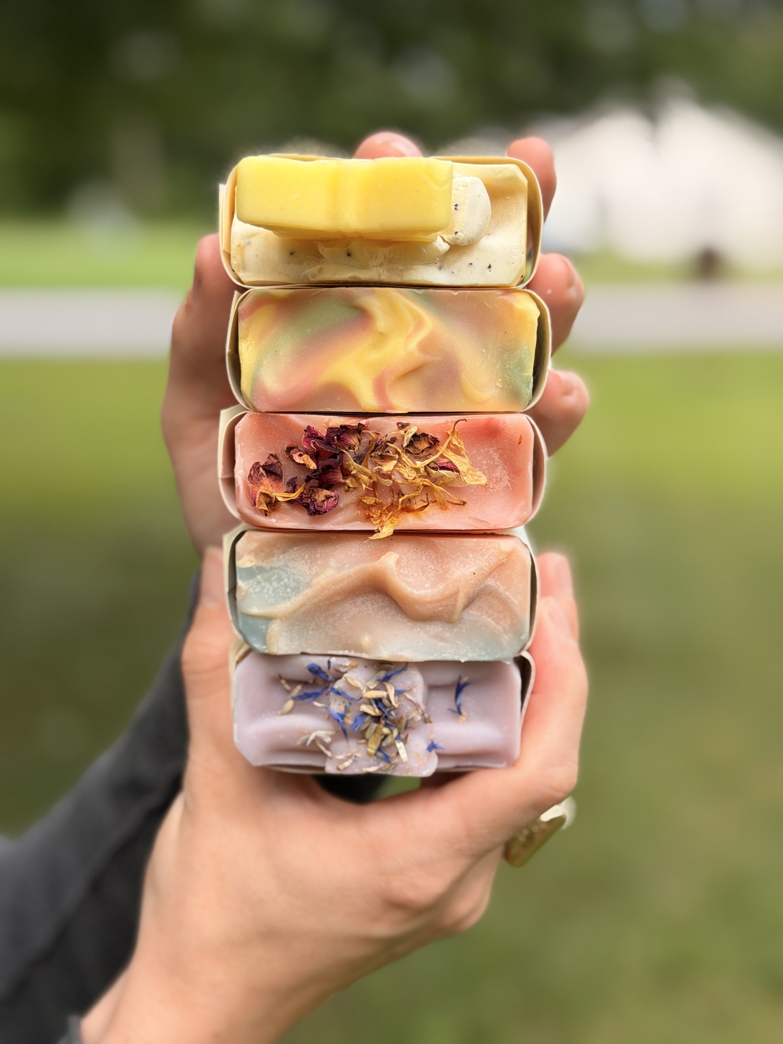A person's hand holding a stack of colorful handmade soap bars with various textures and toppings outdoors.