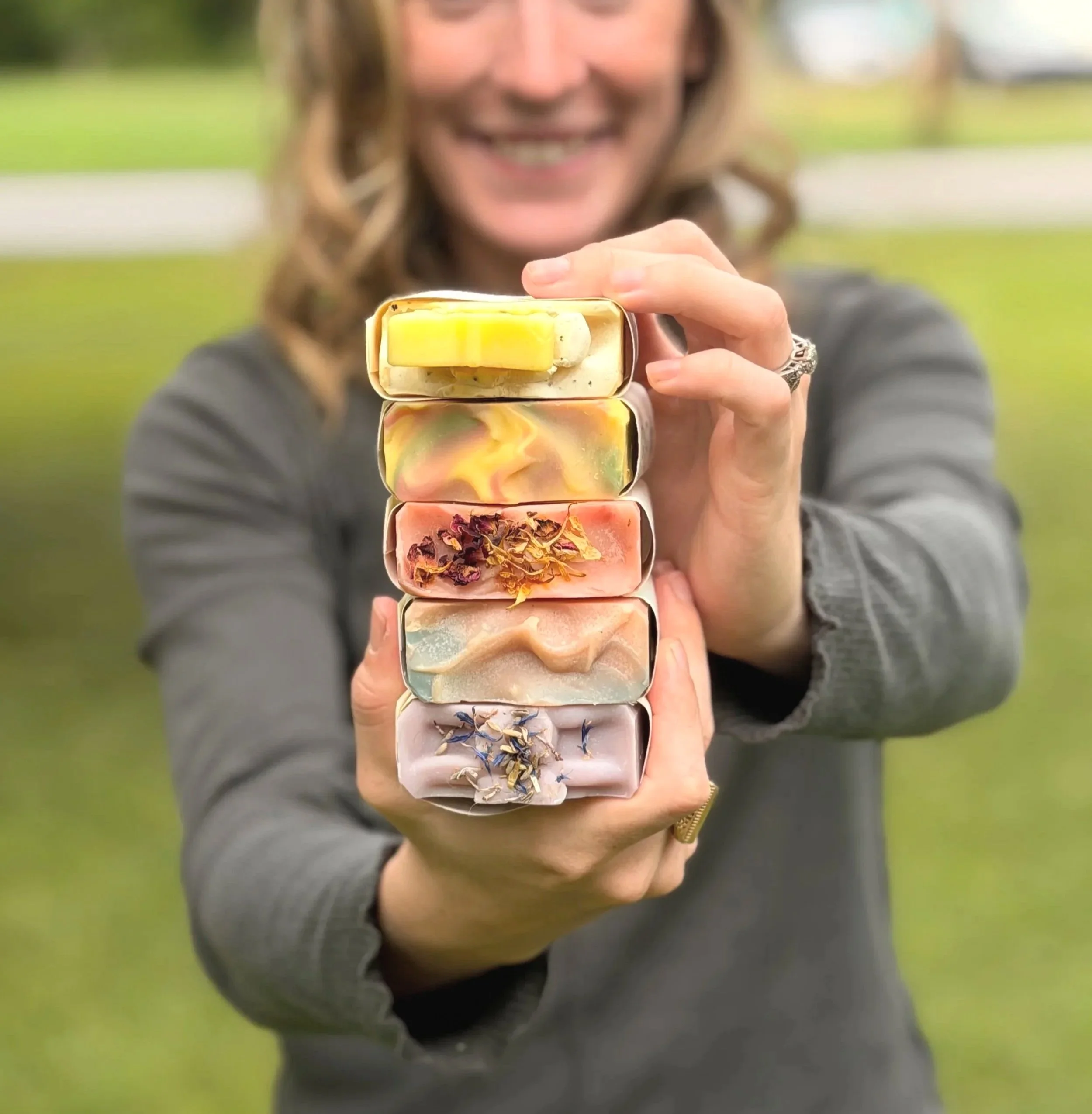 A woman with wavy hair smiling, holding a stack of colorful handmade soap bars outdoors.