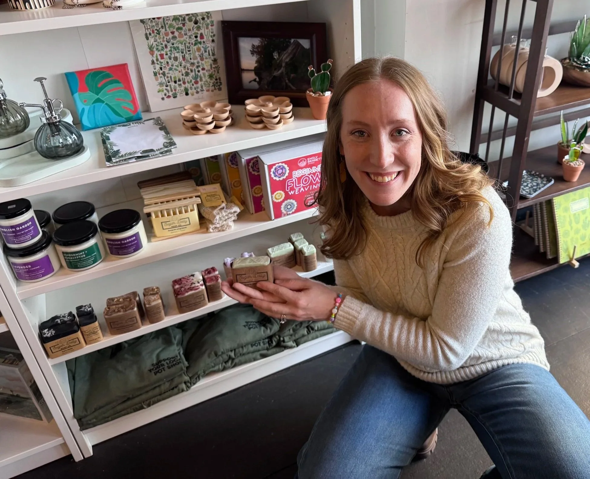 A woman smiling in a store, holding handmade soap bars next to her product display. Behind her, white shelves display various products.