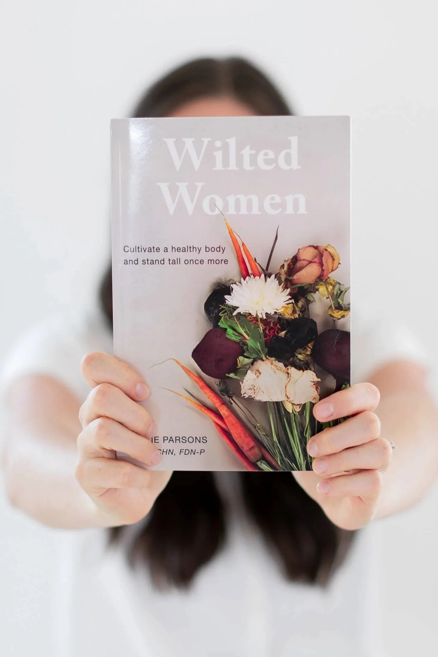 Allie Parsons, FDN-P from Off Beet Holistic FDN, holding a book titled 'Wilted Women' in front of her face. The book cover features images of vegetables and flowers.