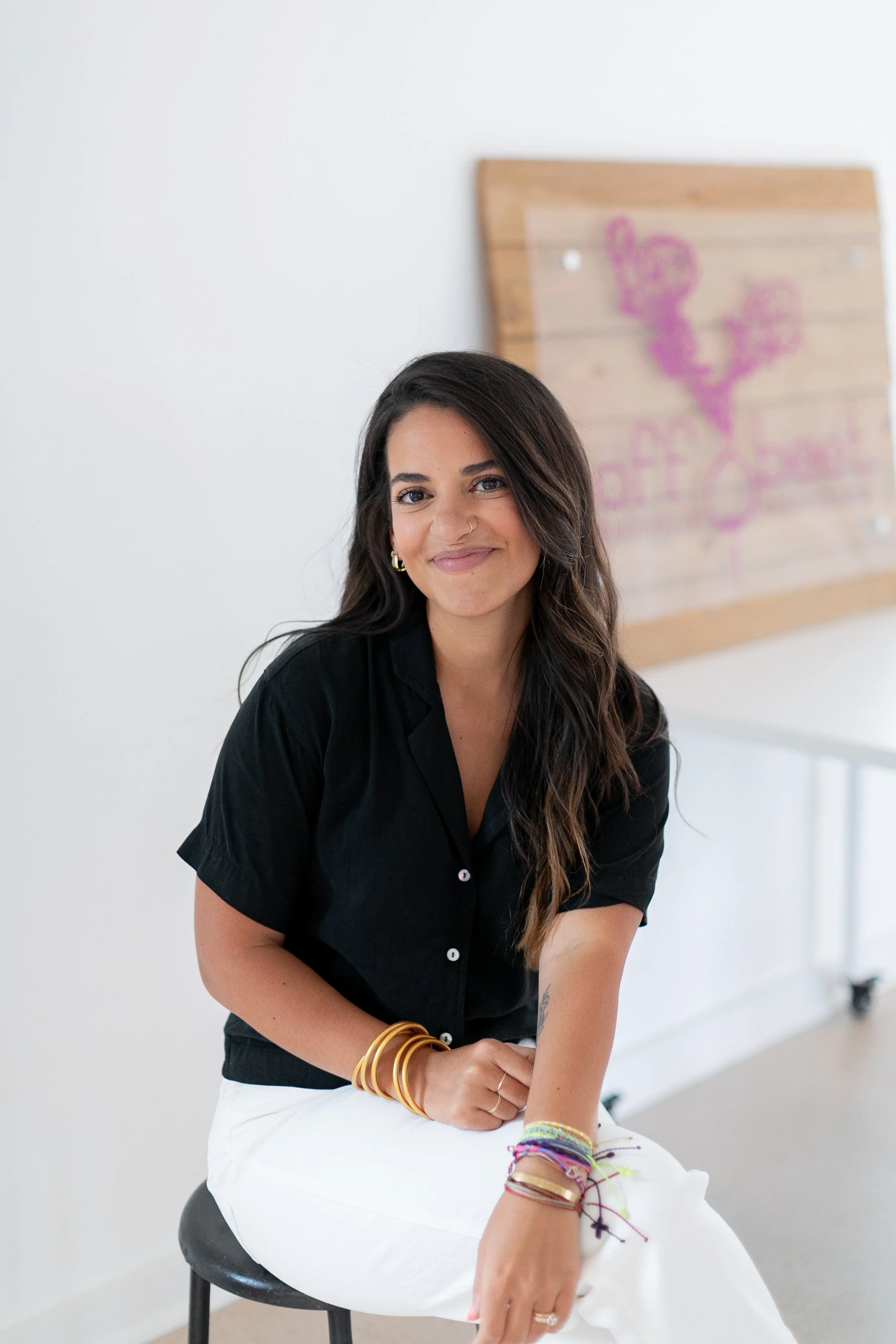A woman sitting on a stool with a smile, wearing a black shirt and white pants, with colorful bracelets on her wrist, in a bright room with a wooden wall art in the background.