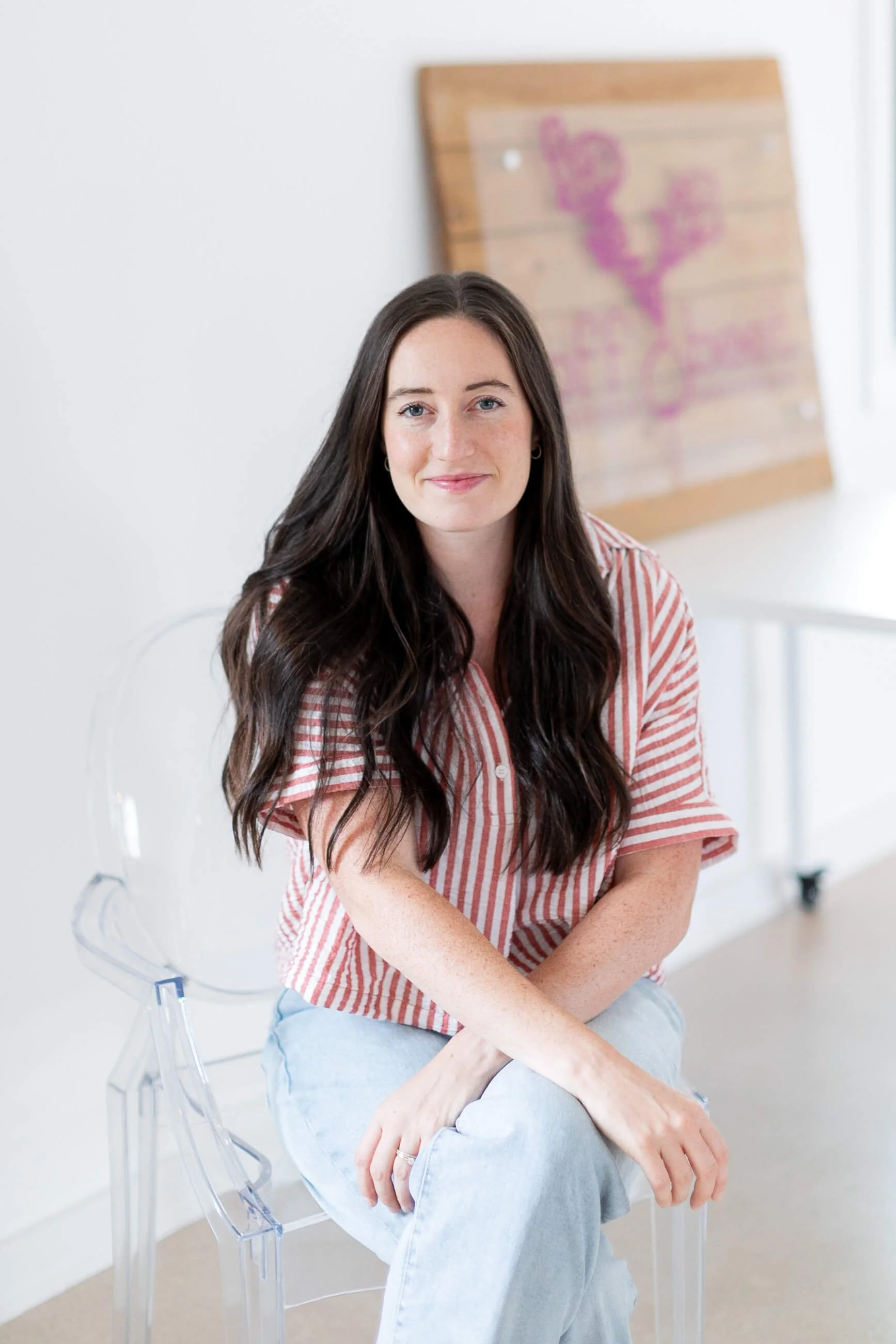 Allie Parsons, FDN-P, from Off Beet Holistic FDN, wearing a pink and white striped shirt and light blue jeans, sitting on a clear acrylic chair in a bright room with a white wall and a wooden art piece with pink and purple paint behind her.