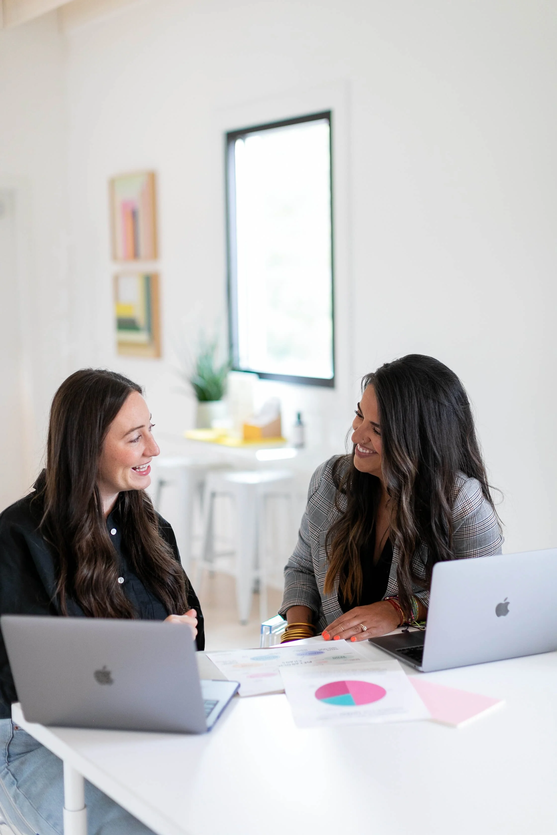 Allie Parsons and Carla Grice with Off Beet Holistic FDN, smiling and talking at a white table with laptops and papers in a bright office space offering functional nutrition services.