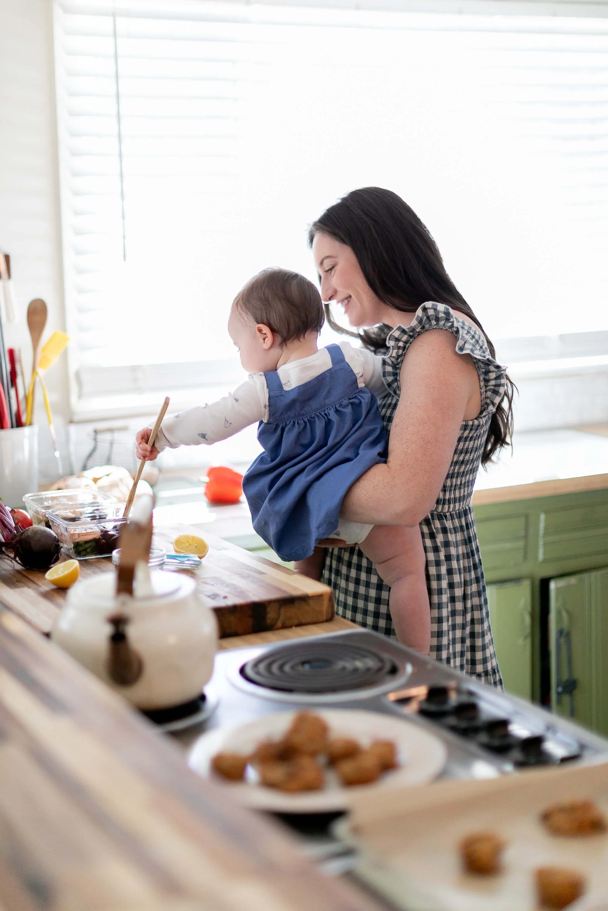 Allie Parsons, FDN-P, holding a young child in a kitchen, smiling as the child reaches for food on the counter, with baked goods and cooking utensils visible.