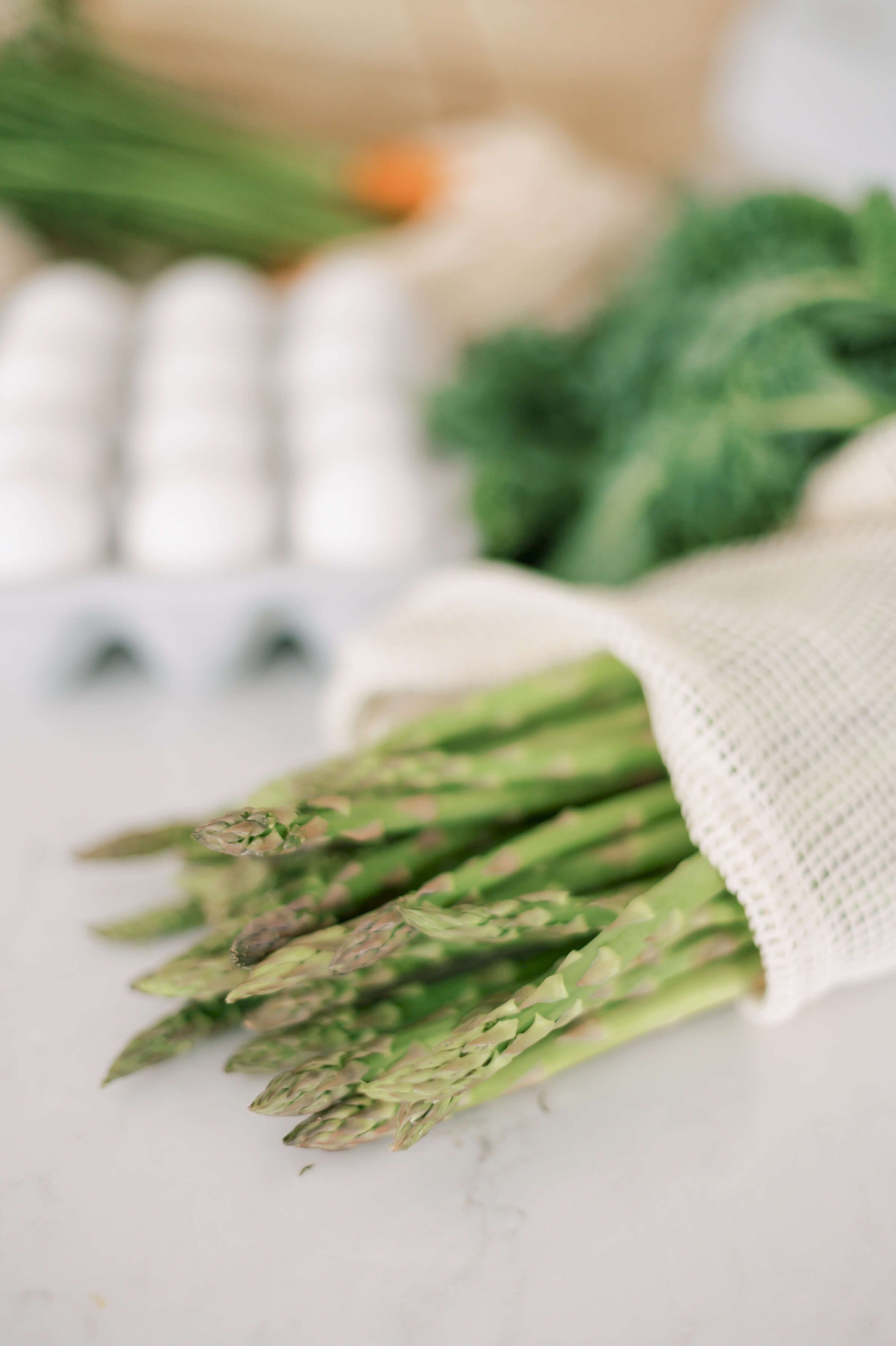 A bunch of fresh green asparagus spears wrapped in a white mesh cloth, lying on a white marble surface at Off Beet Holistic FDN offering functional nutrition services.