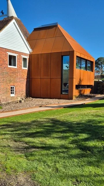 Corten steel-clad extension with an angular roof beside a traditional brick oast house.