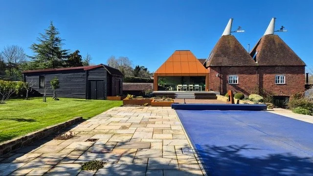 Wide view across a paved garden and pool area towards a Corten steel extension and brick oast house.