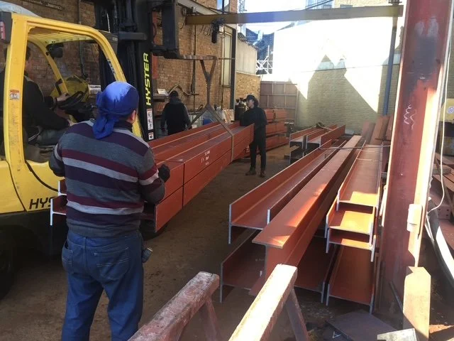 Operatives handling long structural steel beams beside a delivery vehicle in the workshop.