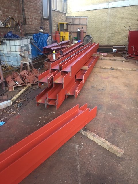 Structural steel sections with red protective primer laid out on the workshop floor.