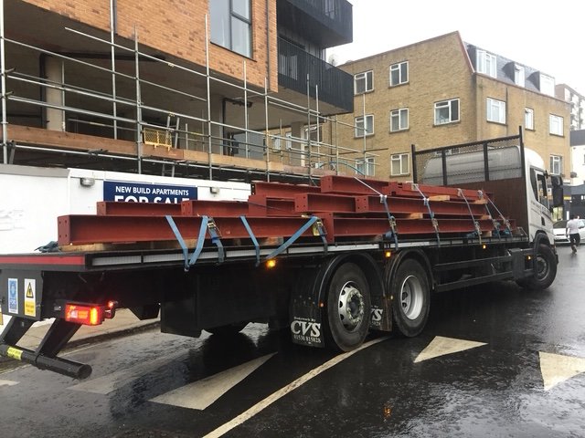 Flatbed lorry delivering red-primed steel beams to an urban construction site.