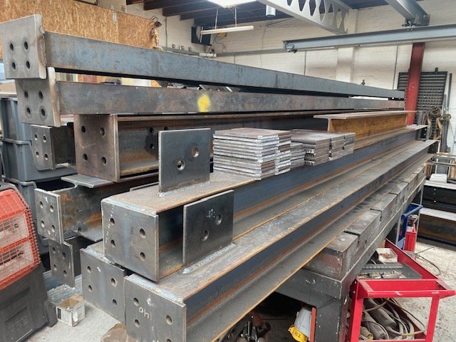 Stacked steel beams with drilled end plates and connection plates ready for fabrication.