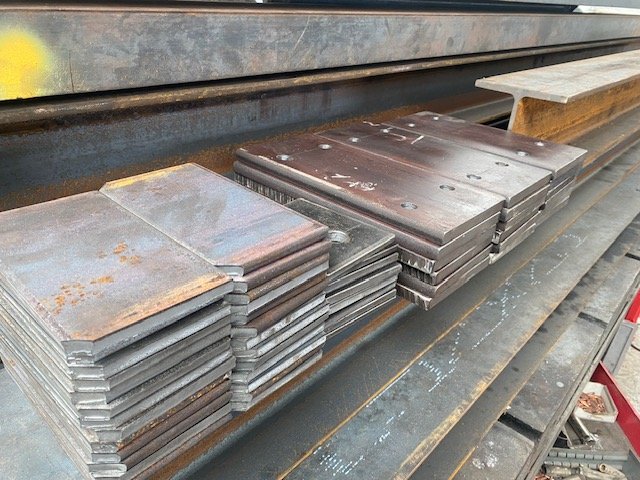 Stacks of drilled steel connection plates beside structural beam sections.