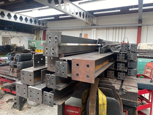 Structural steel beams with bolted connection end plates stacked in the fabrication workshop.