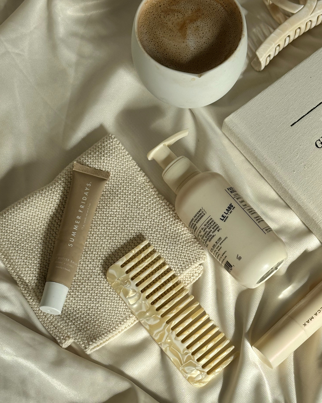 A collection of skincare and beauty items, including a beige tube labeled 'Summer Fridays,' a white cream-colored comb in a marble-patterned case, a bottle of Le Labo product, a textured cloth, a closed book, and a cup of coffee on a satin surface.
