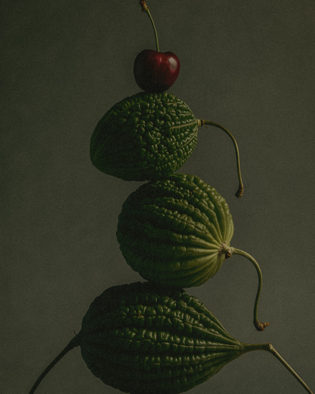 A cherry atop a line of four green bitter melons stacked vertically, with a dark background and soft lighting.