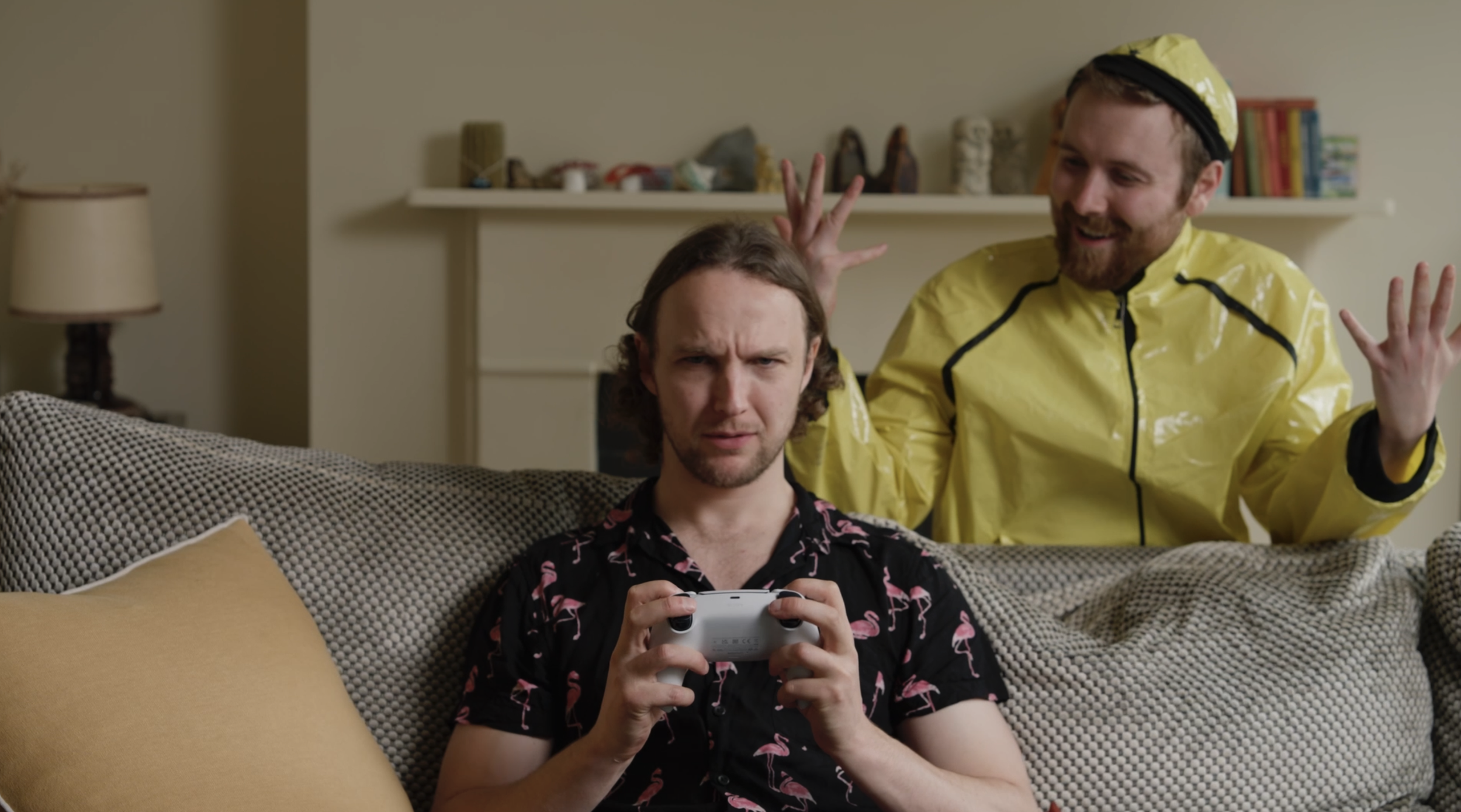 Two men sitting on a couch in a living room, one is looking at a gaming console controller with a confused expression, the other is standing behind him wearing a yellow raincoat and hat, making a humorous gesture.