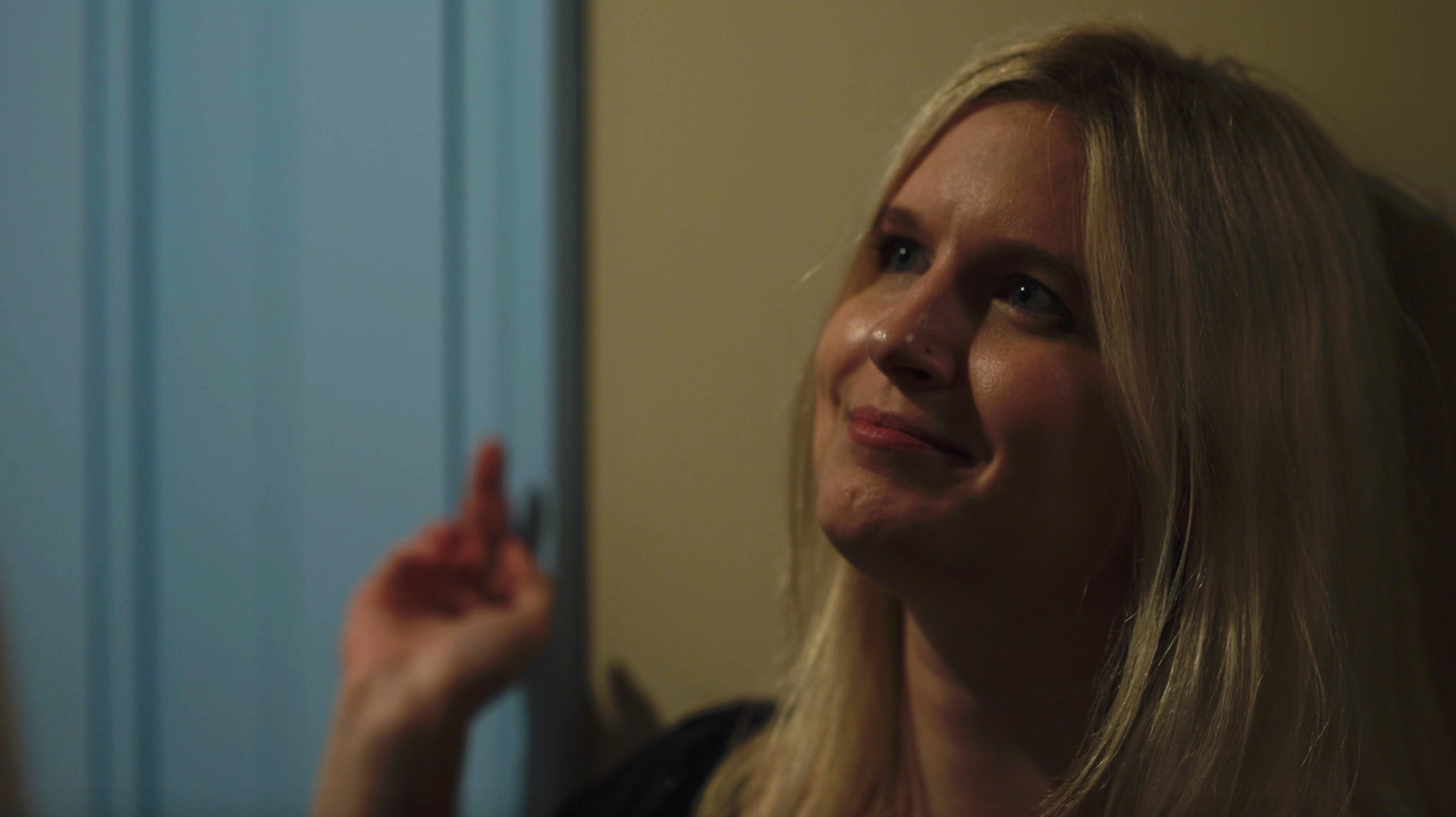 A blonde woman smiling and pointing at something off-camera in a dimly lit room.